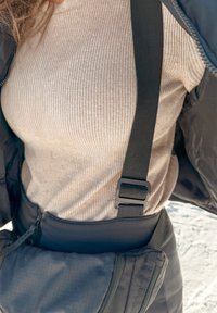 Κοντινό πλάνο ατόμου που φοράει μπεζ πουλόβερ με ραβδώσεις, μαύρο μπουφάν και μαύρο ρυθμιζόμενο λουρί με θήκη φερμουάρ ορατή στη μέση.