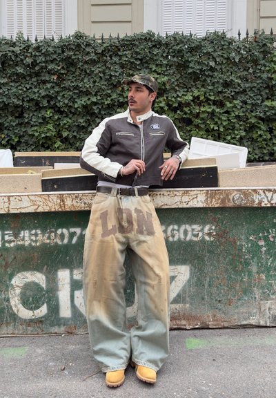 Hombre con gorra de camuflaje, chaqueta en blanco y negro, pantalones beige oversize con estampado "LON" y botas amarillas, apoyado en un contenedor verde al aire libre.