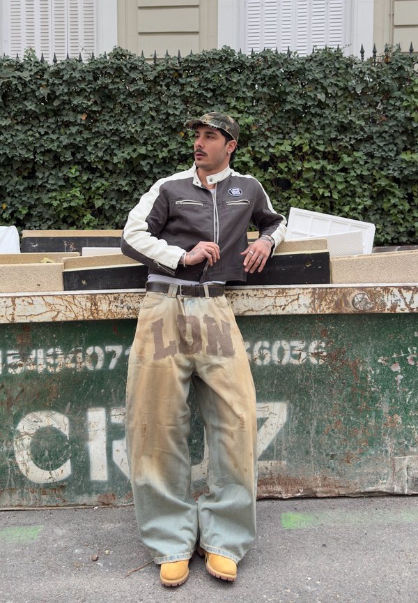 Άνδρας με καπέλο καμουφλάζ, ασπρόμαυρο μπουφάν, φαρδιά μπεζ παντελόνια με τύπωμα "LON" και κίτρινα μποτάκια στηρίζεται σε έναν πράσινο κάδο απορριμμάτων σε εξωτερικό χώρο.