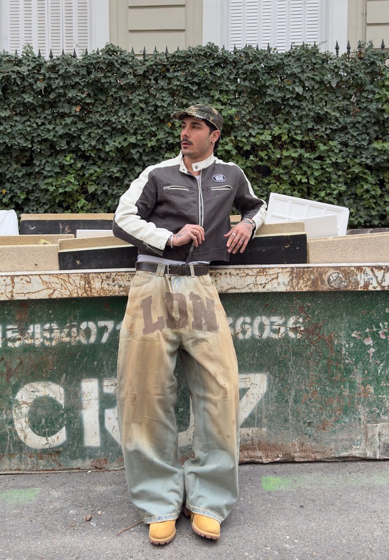 Moški z maskirno kapo, črno-belo jakno, prevelikimi bež hlačami z napisom "LON" in rumenimi škornji se naslanja na zeleno smetnjak na prostem.