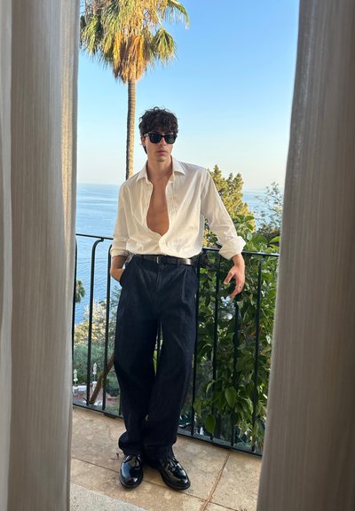 Camisa blanca de botones, abierta en el cuello, combinada con pantalones de mezclilla oscuros y zapatos negros. El fondo presenta plantas y una vista al mar.