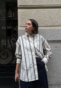 Donna con capelli corti e scuri che indossa una camicia bianca a righe larga e pantaloni scuri, in piedi contro un muro di pietra strutturato con una mano in tasca.