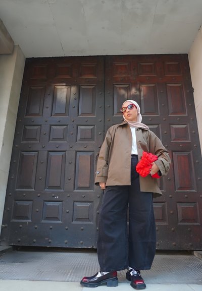 Chaqueta marrón, camisa blanca, pantalones de pierna ancha negros, accesorio rojo con volantes, gafas de sol con tintado rojo, zapatos de plataforma negros, con puertas de madera de fondo.