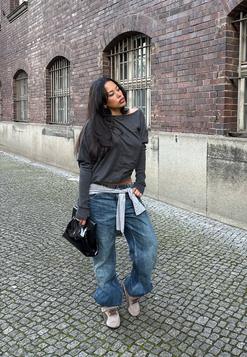 Langärmliges dunkelgraues Oberteil, hellgrauer gebundener Blazer, lockere blaue Jeans und beiger Sneaker. Schwarze Handtasche, gepflasterte Straße aus Naturstein. Ziegelmauer im Hintergrund.