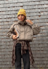 Veste beige courte avec col montant, manches froncées et texture mate. Associée à un jean foncé et une chemise à carreaux nouée. Bonnet en tricot jaune porté.