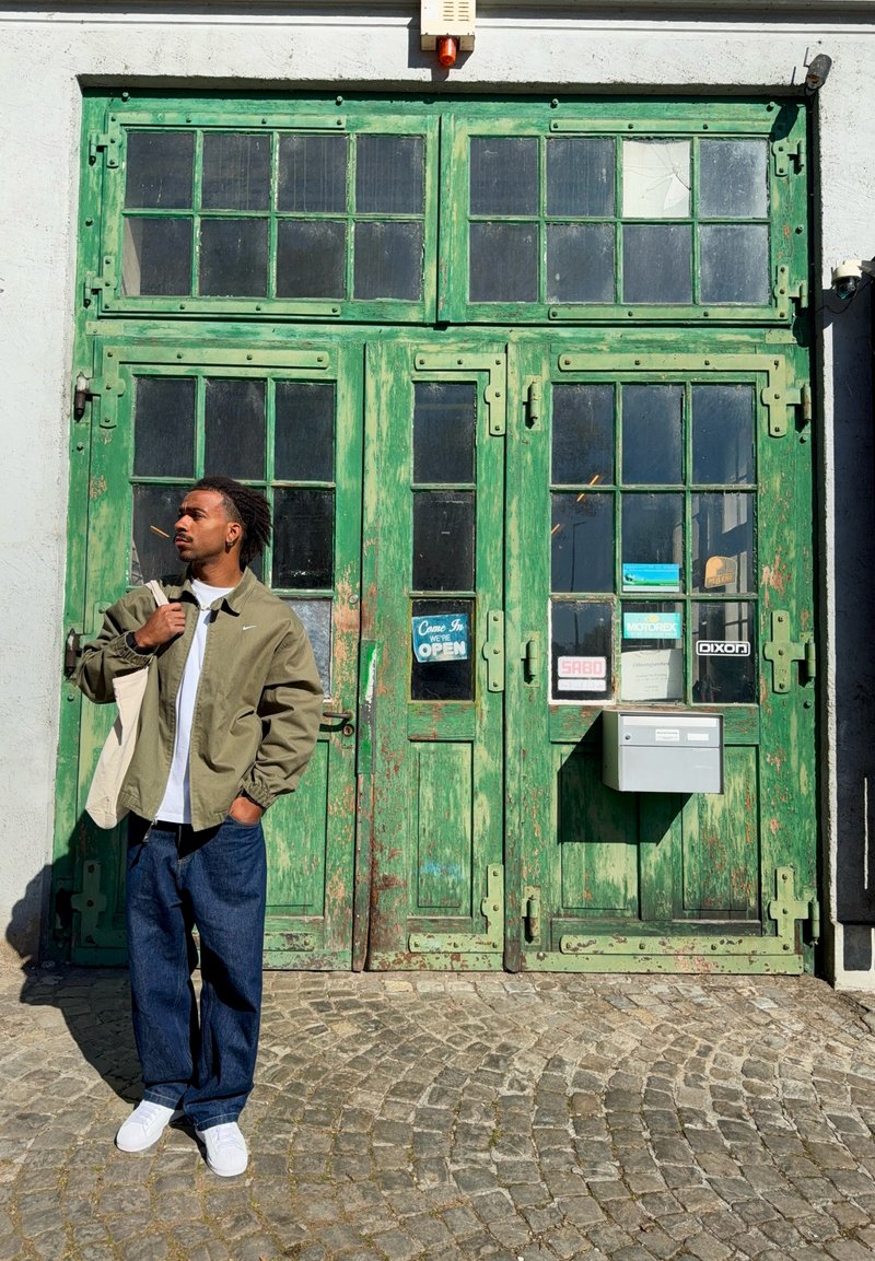 Man in groene jas en jeans staat met tas over de schouder voor versleten groene houten deuren met glas en een bordje "open".