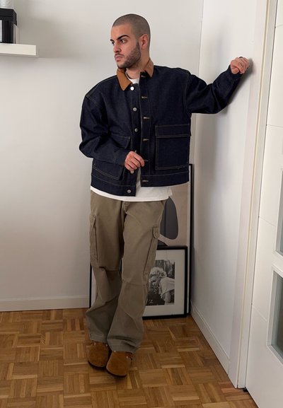 Chaqueta de denim oscura con un cuello beige, que presenta bolsillos frontales y botones; combinada con pantalones cargo beige y calzado marrón.