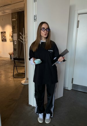 Juoda didelė marškinė su balta logo, juodos sportinės kelnės su baltais dryžiais, baltos sportinės bateliai, laikantys šviesiai mėlyną puodelį ir planšetę.