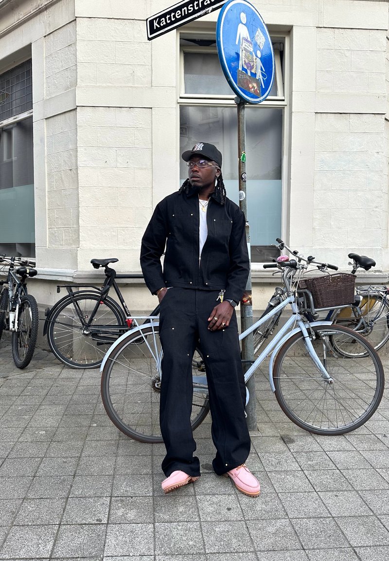 Svart denimjacka och byxor med gyllene detaljer, tillsammans med rosa skor. Bakgrunden har cyklar och ett gatukänt.