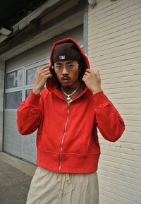 Sweatshirt vermelho com fecho zip e textura suave, apresentando um bolso canguru e capuz ajustável com cordão, usado sobre cal�ças beje claras.