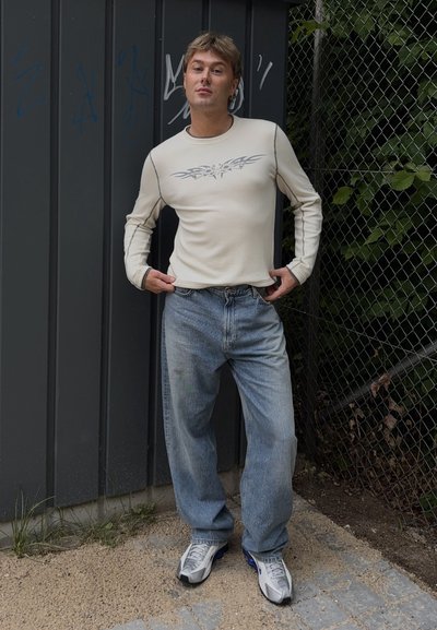 Camisa de manga larga de color beige claro con diseño gráfico, combinada con jeans relajados de color azul claro y zapatillas grises. De pie contra una valla.