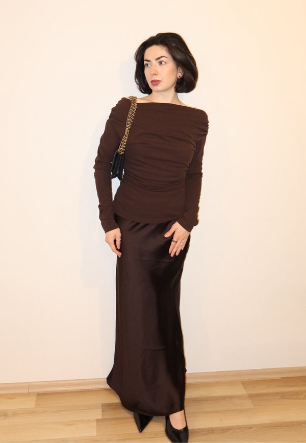 Femme aux cheveux courts et foncés portant une longue robe marron et des talons noirs, tenant un sac noir avec une chaîne dorée, debout contre un mur beige.