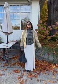 Ljusbeige zip-up jacka över en svart och grå randig polotröja, parat med en vit maxikjol. Håller en mörk väska; höstlöv omger.