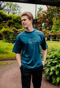 Turkusowa koszulka z krótkim rękawem z białym napisem graficznym "Cape Cod Sailing Club" oraz wzorem łodzi. Noszona z czarnymi spodniami.