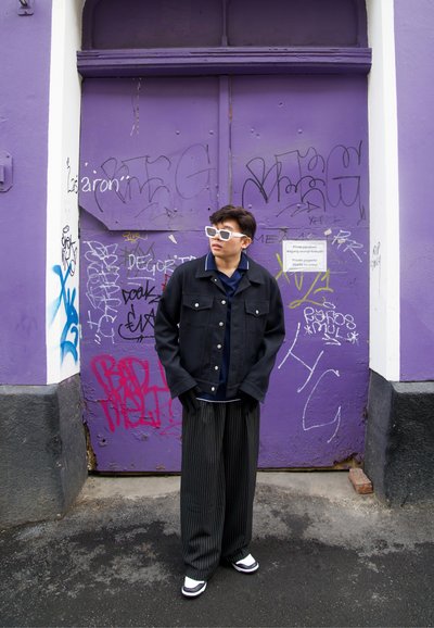 Chaqueta de mezclilla negra sobre una camisa azul marino, pantalones anchos a rayas y zapatillas blancas. Grandes gafas de sol blancas. Pared de graffiti púrpura de fondo.