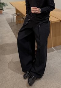 Pantalon noir à jambes larges en tissu texturé, avec de gros boutons latéraux et une coupe décontractée. Associé à des chaussures noires brillantes.