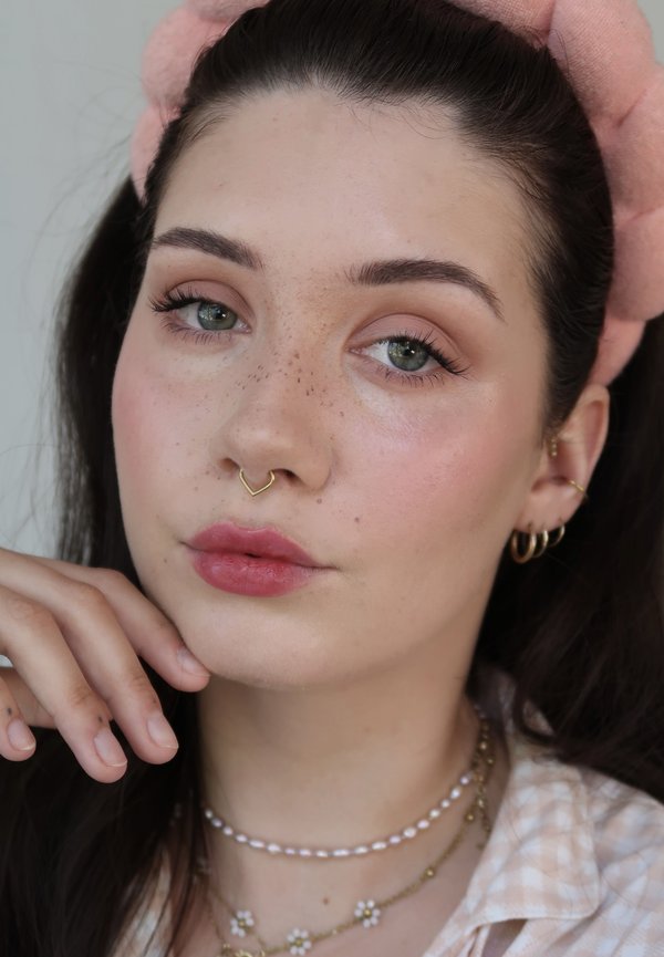 Jonge vrouw met lichte sproeten die een roze haarband, gouden septumring, gouden oorringen, parel- en bloemkettingen draagt, en haar kin op haar hand laat rusten.