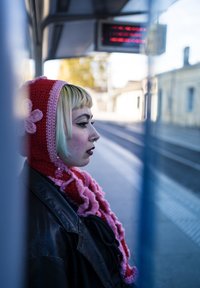 Strikket rød hette med rosa aksenter og blomsterdetalj, kombinert med en svart skinnjakke. Personen har kort, lysegrønt hår og øredobber.