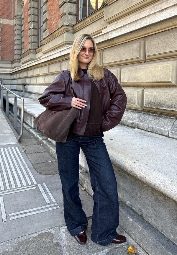 Smeđa kožna bomber jakna nošena preko tamnog džempera, uparena s tamnim trapericama širokih nogavica i smeđim ravnim cipelama. Nose se smeđa antilop torba na ramenu.