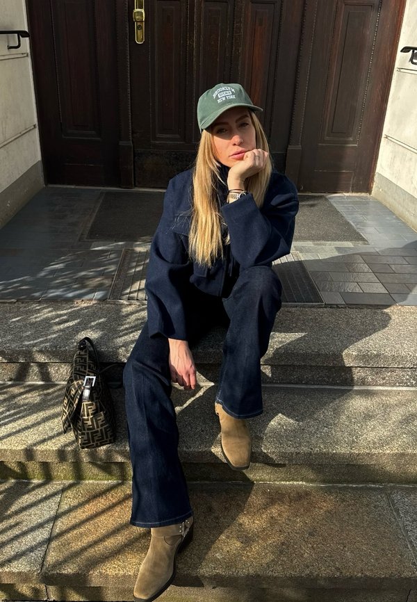 Jonge vrouw in marineblauwe outfit en groene pet zittend op stenen trappen in het zonlicht, met een handtas met patroon naast haar.