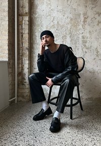 Melnas ribotas garām piedurknēm krekls, ādas jakas, melnas mākslīgās ādas bikses, baltas zeķes, melni loaferi ar ķēdes detaļu, sēž uz krēsla.