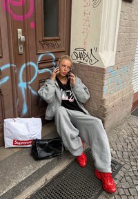 Grå oversized hoodie og sweatpants, røde sneakers, sort håndtaske med kædestropp og hvid indkøbspose med rødt logo foran en murstensvæg.