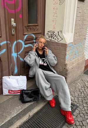 Sudadera gris de gran tamaño y pantalones de chándal, zapatillas rojas, bolso negro con cadena, y bolsa de compras blanca con logotipo rojo contra una pared de ladrillos.