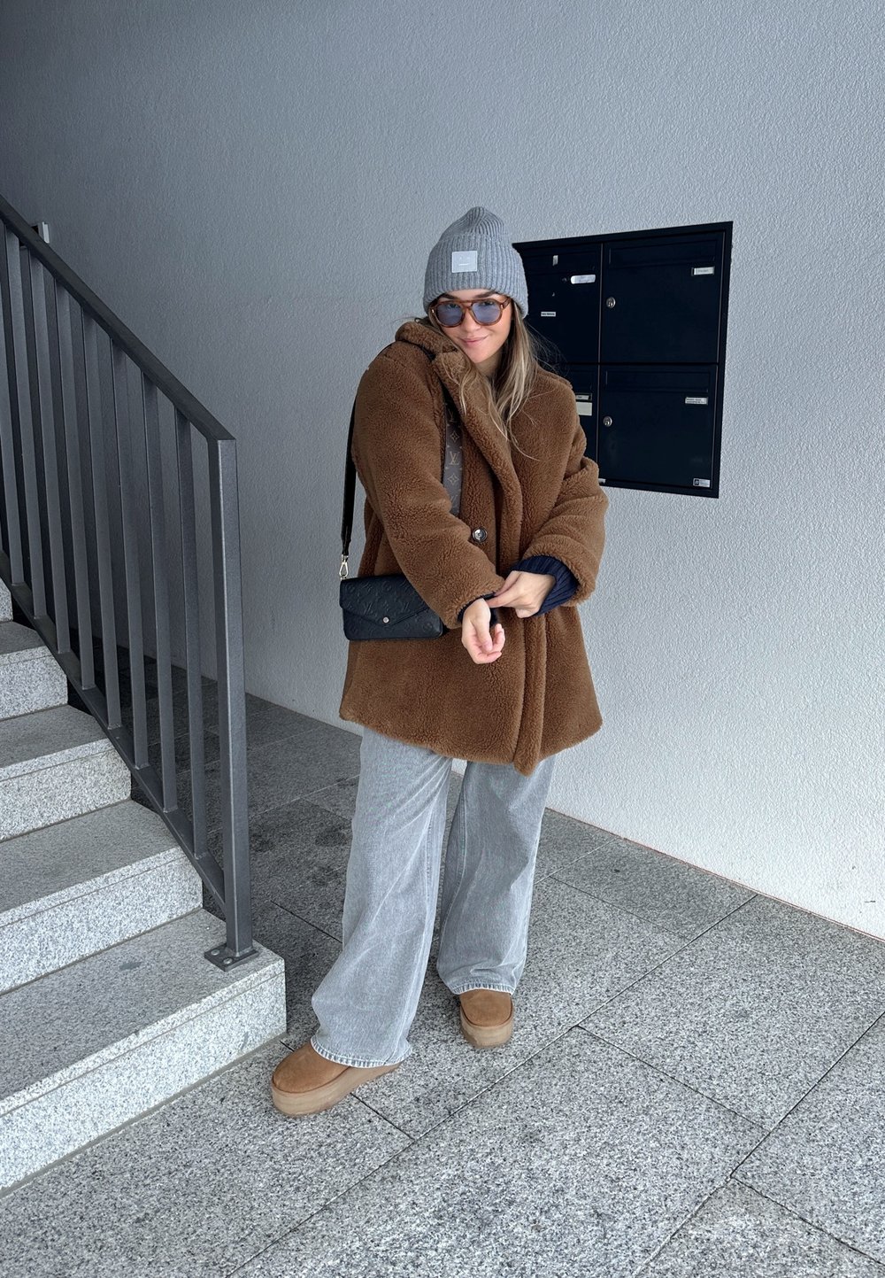 Brūns mākslīgās kažokādas mētelis, pelēks beanie, saulesbrilles un melna plecu soma; pelēki platās kājas bikses un brūnas platformas kurpes; stāv blakus kāpnēm.