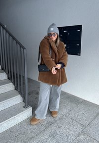 Brūns mākslīgās kažokādas mētelis, pelēks beanie, saulesbrilles un melna plecu soma; pelēki platās kājas bikses un brūnas platformas kurpes; stāv blakus kāpnēm.