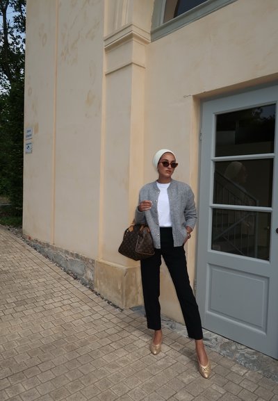 Cardigan de punto gris, camiseta blanca, pantalones negros, bailarina dorada. Bolso marrón con estampado. De pie contra una pared beige cerca de una puerta gris.