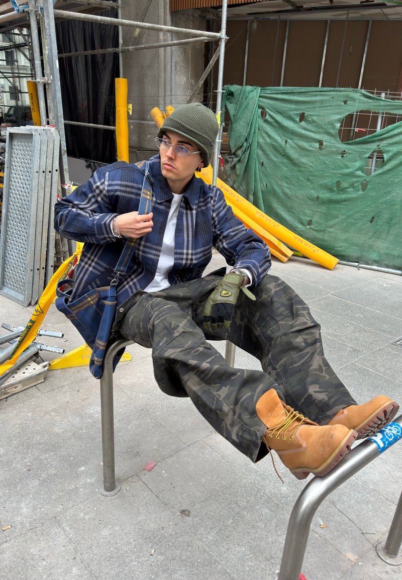 Jeune personne en pantalon camouflage et bottes marron clair, allongée sur une rambarde métallique au milieu d'un échafaudage de construction et d'une bâche verte déchirée.