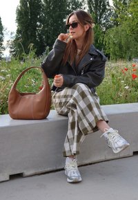 Καφέ δερμάτινη τσάντα, μαύρο δερμάτινο τζάκετ, πράσινο και λευκό καρό παντελόνι, ασημένια αθλητικά παπούτσια, καθισμένη σε μια τσιμεντένια παγκάκι σε μια ανθισμένη διακόσμηση.