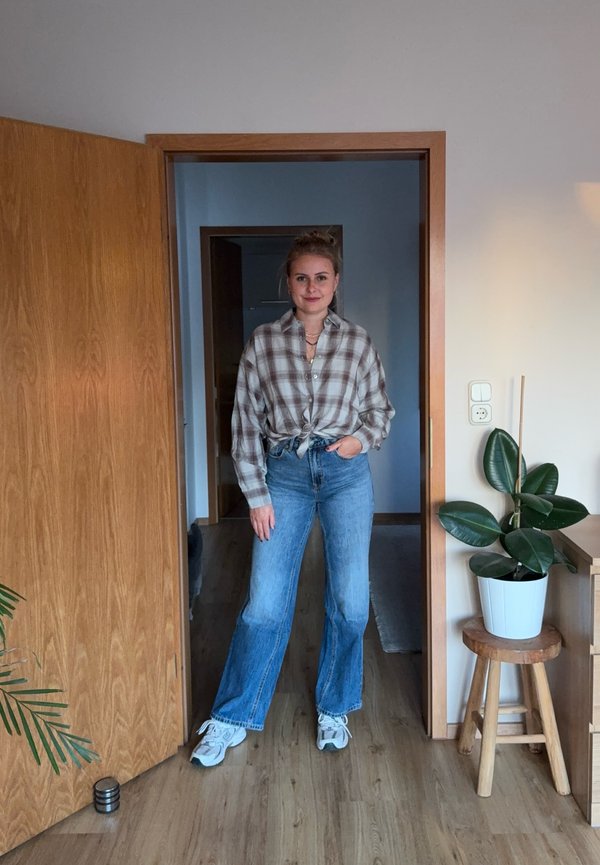 Une femme se tient dans l’embrasure d’une porte, vêtue d’une chemise à carreaux, d’un jean large et de baskets ; à côté d’elle, une plante en pot est posée sur un tabouret en bois.