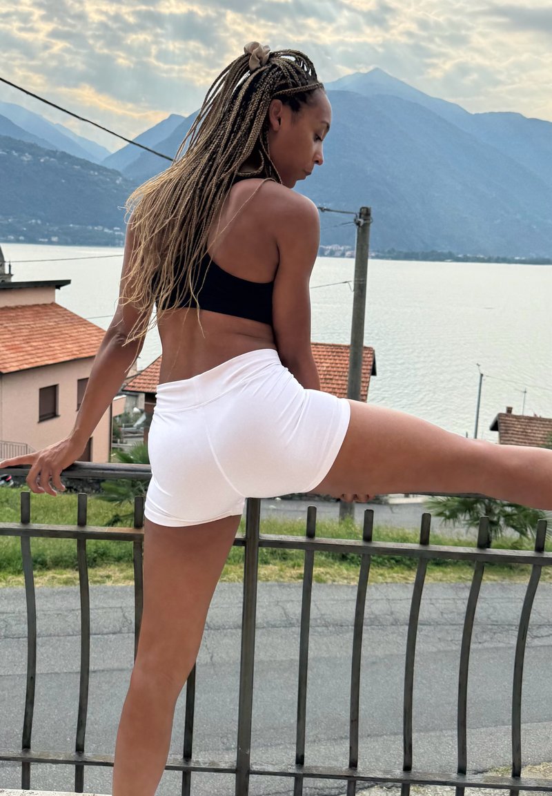 Woman in black sports bra and white shorts stretching leg on railing with lake, houses, and mountains in the background under cloudy sky.