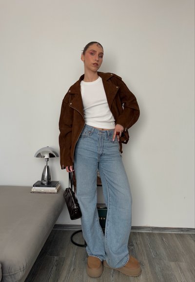 Chaqueta de motociclista de ante marrón, camiseta blanca ajustada, jeans de pierna ancha azul claro, zapatos de plataforma color beige y un pequeño bolso negro.