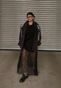 Μαύρο φούτερ με κουκούλα και λευκές ρίγες, επενδυμένο πάνω από ένα μαύρο φόρεμα με υφή δαντέλας. Μαύρα, χοντρά παπούτσια και γυαλιά ηλίου ολοκληρώνουν το λουκ.