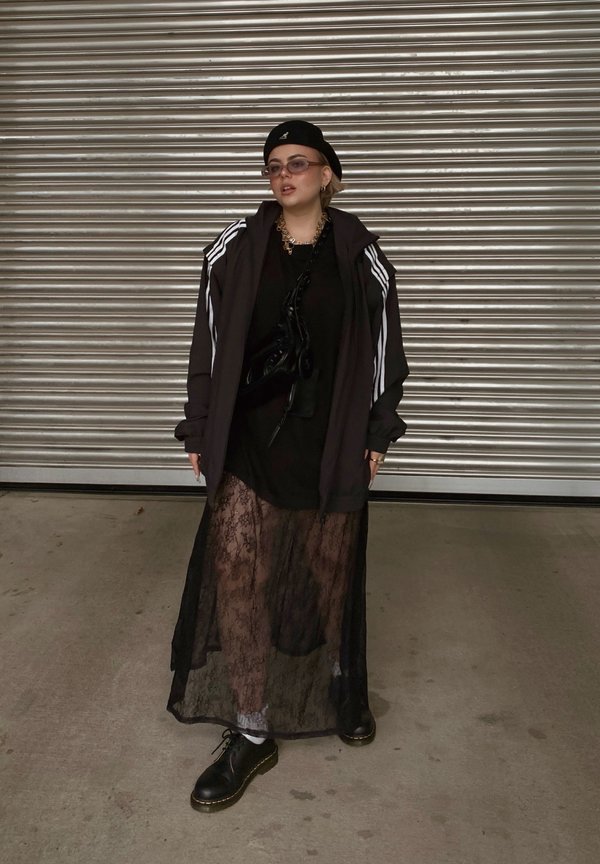 Zwarte hoodie met witte strepen, gelaagd over een zwarte jurk met kanttextuur. Zwarte, chunky schoenen en een zonnebril maken de look compleet.