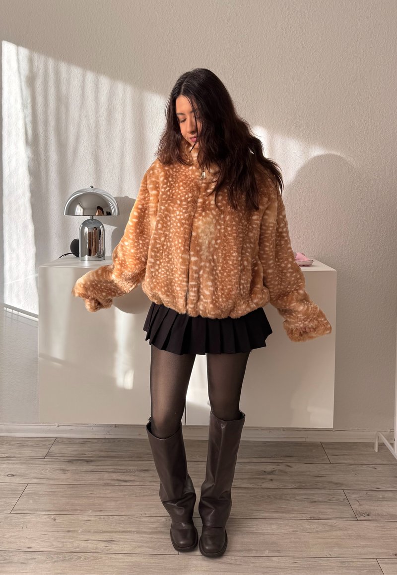 Giovane donna che indossa una giacca in pelliccia sintetica marrone maculata, gonna plissettata nera, calze nere e stivali alti marrone scuro, in piedi all'interno.