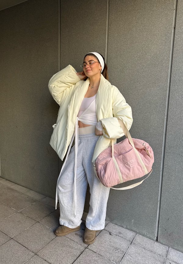 Kvinde iført cremefarvet dynejakke, hvid crop top, grå joggingbukser og støvler, der holder en pink sportstaske og bærer briller samt et hvidt pandebånd.