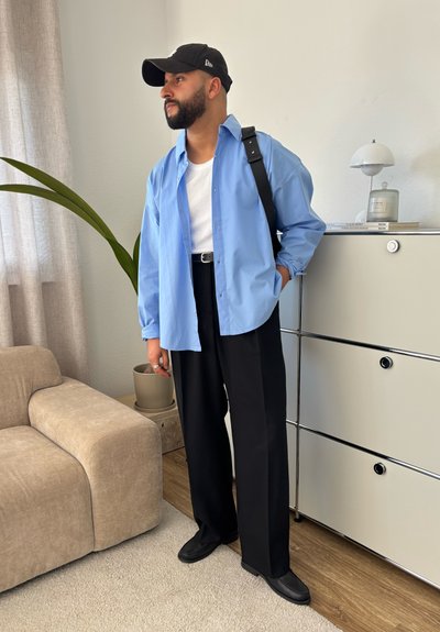 Camisa de botones azul claro sobre una camiseta blanca, pantalones anchos negros y zapatos negros. Los accesorios incluyen una gorra negra y un cinturón.