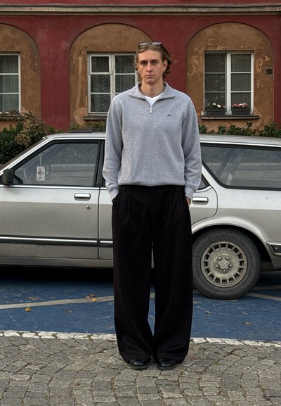 Sudadera gris con cremallera y logo, pantalones anchos negros y zapatos negros. De fondo, un coche plateado aparcado y un pavimento de piedra texturizado.