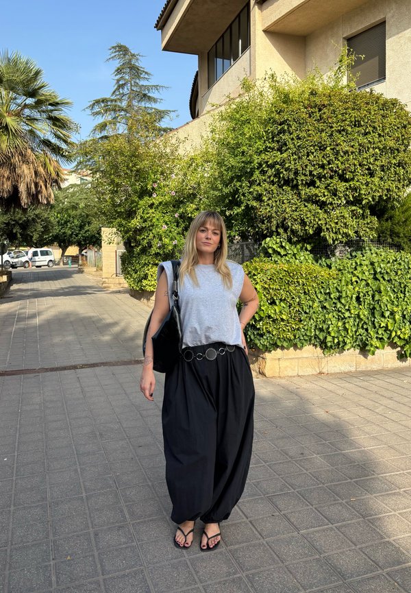 Young woman standing on a paved sidewalk near green bushes and trees, wearing a gray sleeveless top and loose black pants.