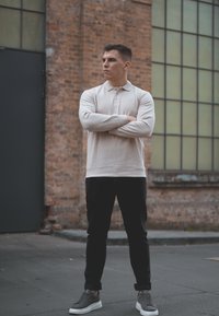 Ανοιχτό μπεζ μακρυμάνικο polo με ριγωτή υφή, σκούρο παντελόνι και γκρι αθλητικά παπούτσια. Το μοντέλο στέκεται με αυτοπεποίθηση με τα χέρια σταυρωμένα.