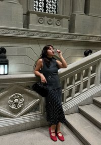 Femme en haut noir sans manches et jupe à pois ajuste ses lunettes en s'appuyant sur une rampe en pierre ornée près d'un escalier dans un intérieur grandiose.