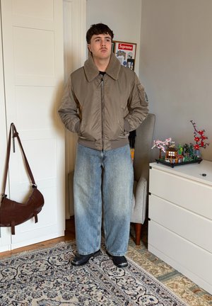 Joven de pie en el interior con una chaqueta marrón con cuello de piel, jeans anchos azul claro, zapatos negros, junto a una silla y una cómoda blanca con decoraciones.