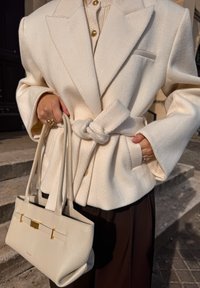 Krémový blejzer so vzorom rybej kosti a opaskom, nosený cez svetlý top. Svetlá kabelka so zlatým detailom držená v ruke.