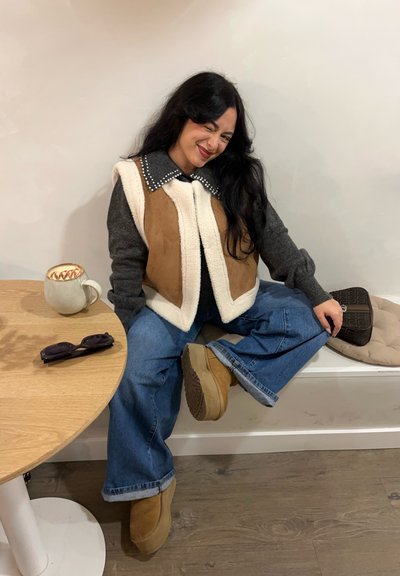 Chaleco de color marrón y crema con forro de polar, suéter gris, vaqueros de pierna ancha azules, botas marrones, taza redonda y gafas de sol negras sobre una mesa.