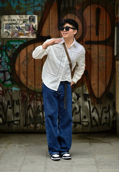Camisa blanca de manga larga con costuras en contraste, vaqueros anchos azules, zapatillas de deporte en blanco y negro, bolso cruzado marrón. Fondo urbano con grafitis.