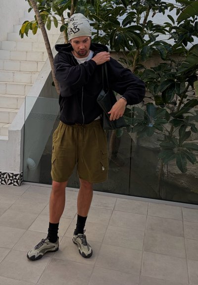 Joven con pantalones cortos beige, sudadera negra con capucha, bandana blanca y zapatillas ajustando una bolsa negra de hombro en un área exterior con baldosas y plantas.