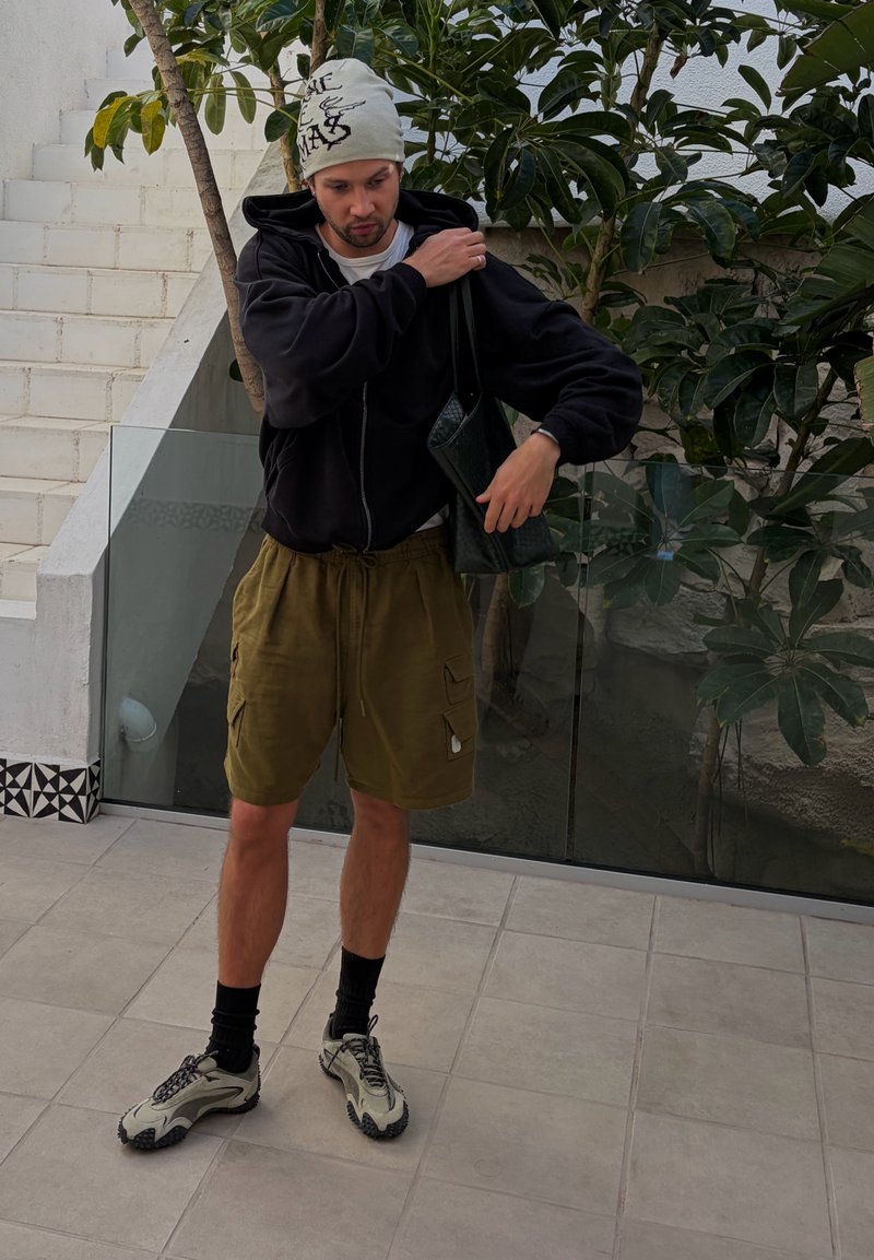 Jeune homme portant un short beige, un sweat à capuche noir, un bandana blanc et des baskets, ajustant un sac à bandoulière noir dans un espace extérieur carrelé avec des plantes.